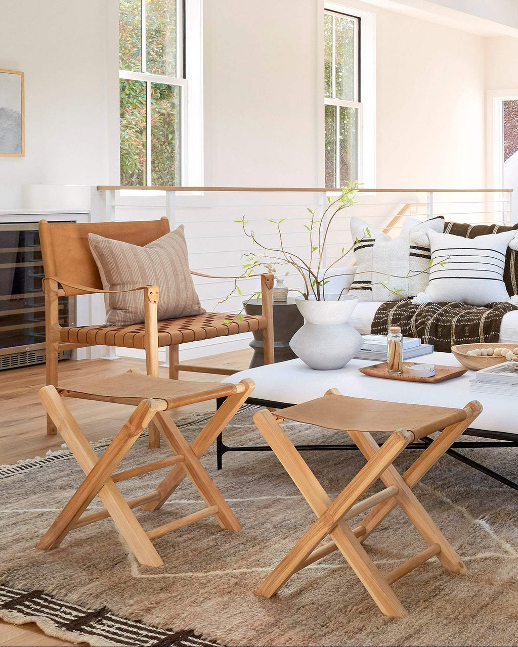 Two light natural wood folding director-style leather folding stools shown in a bright modern living room beside a white coffee table and sofa.