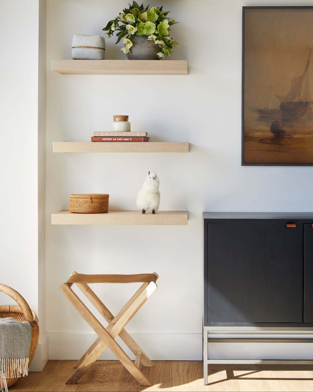 A styled interior scene showing a set of five light natural wood floating shelves mounted on a white wall with a leather folding stool beneath.