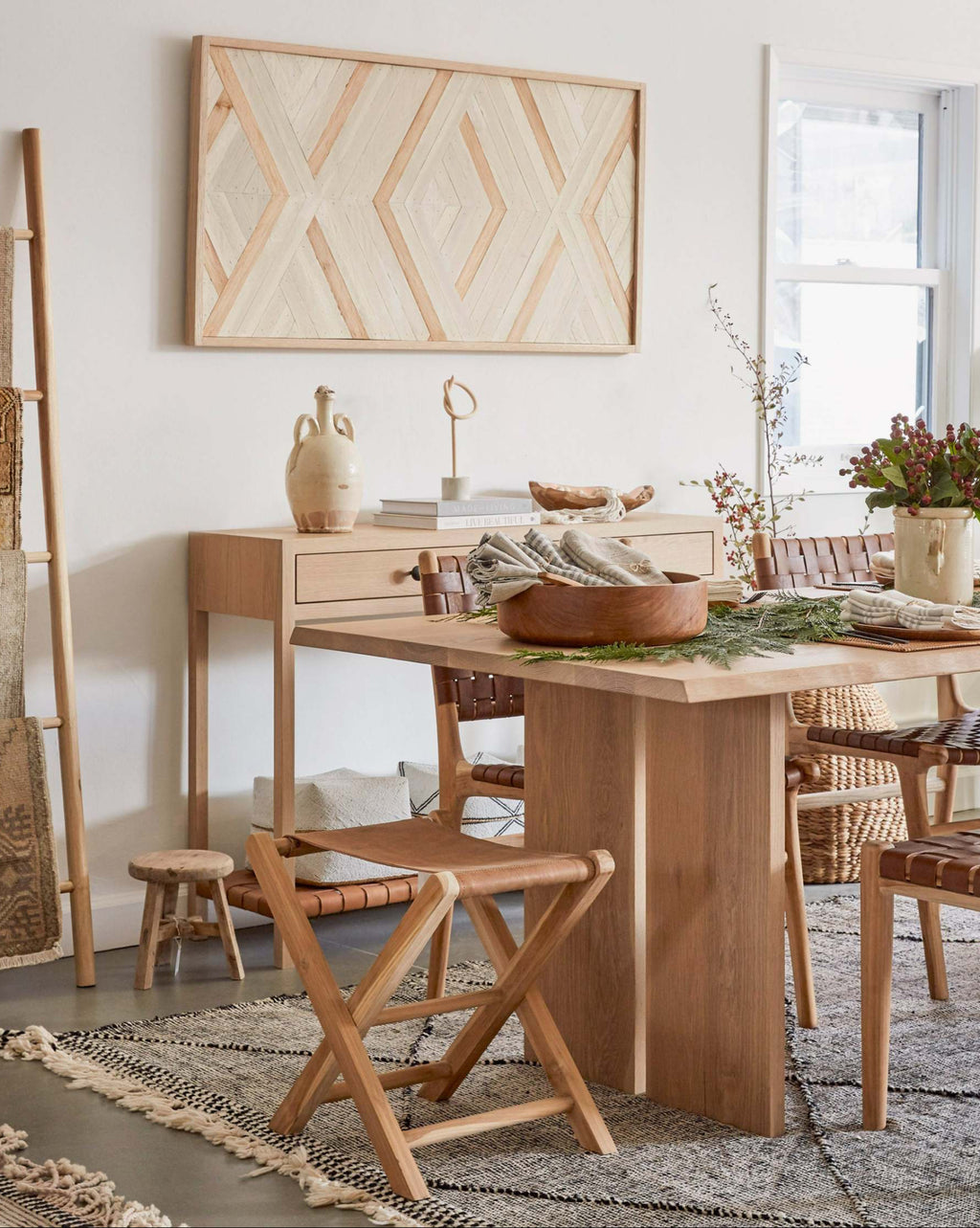 A styled dining room scene featuring a light natural wood dining table and a leather folding stool, a sideboard with ceramic vases, a geometric framed wall art piece, and a patterned rug in a bright, minimalist interior.