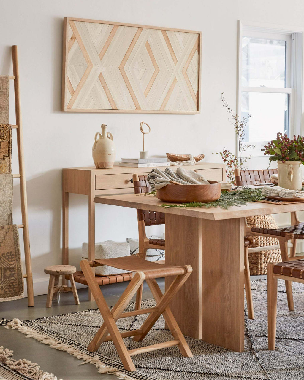 A styled dining room scene featuring a light natural wood dining table and a leather folding stool, a sideboard with ceramic vases, a geometric framed wall art piece, and a patterned rug in a bright, minimalist interior.