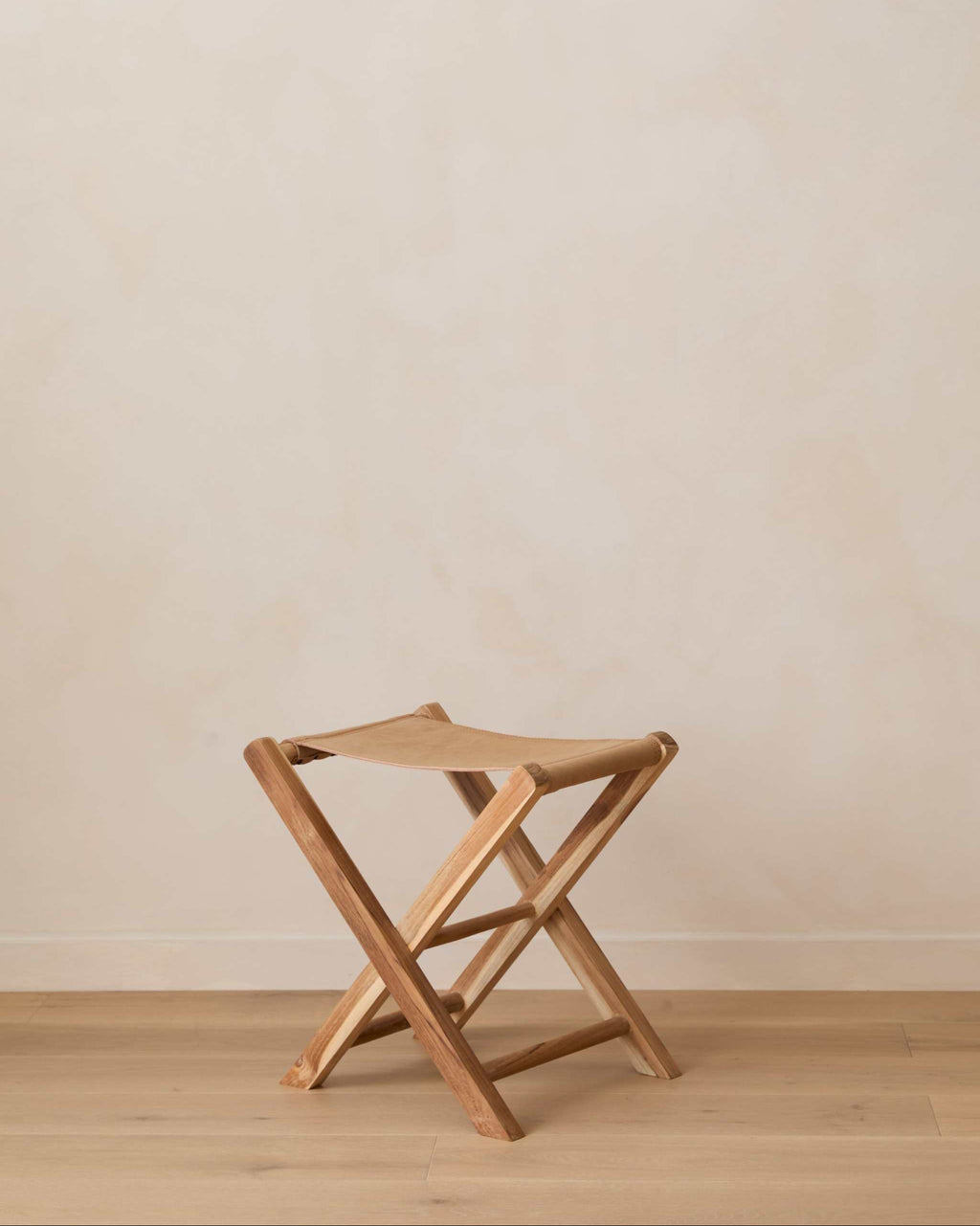 A small leather folding stool shown on a light wood floor against a plain beige wall.