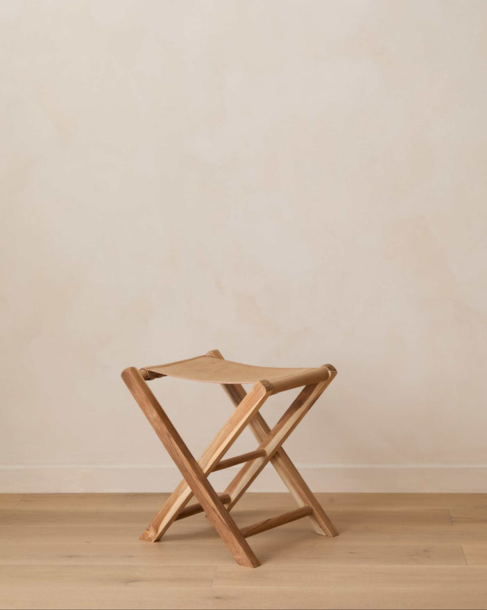 A small leather folding stool shown on a light wood floor against a plain beige wall.