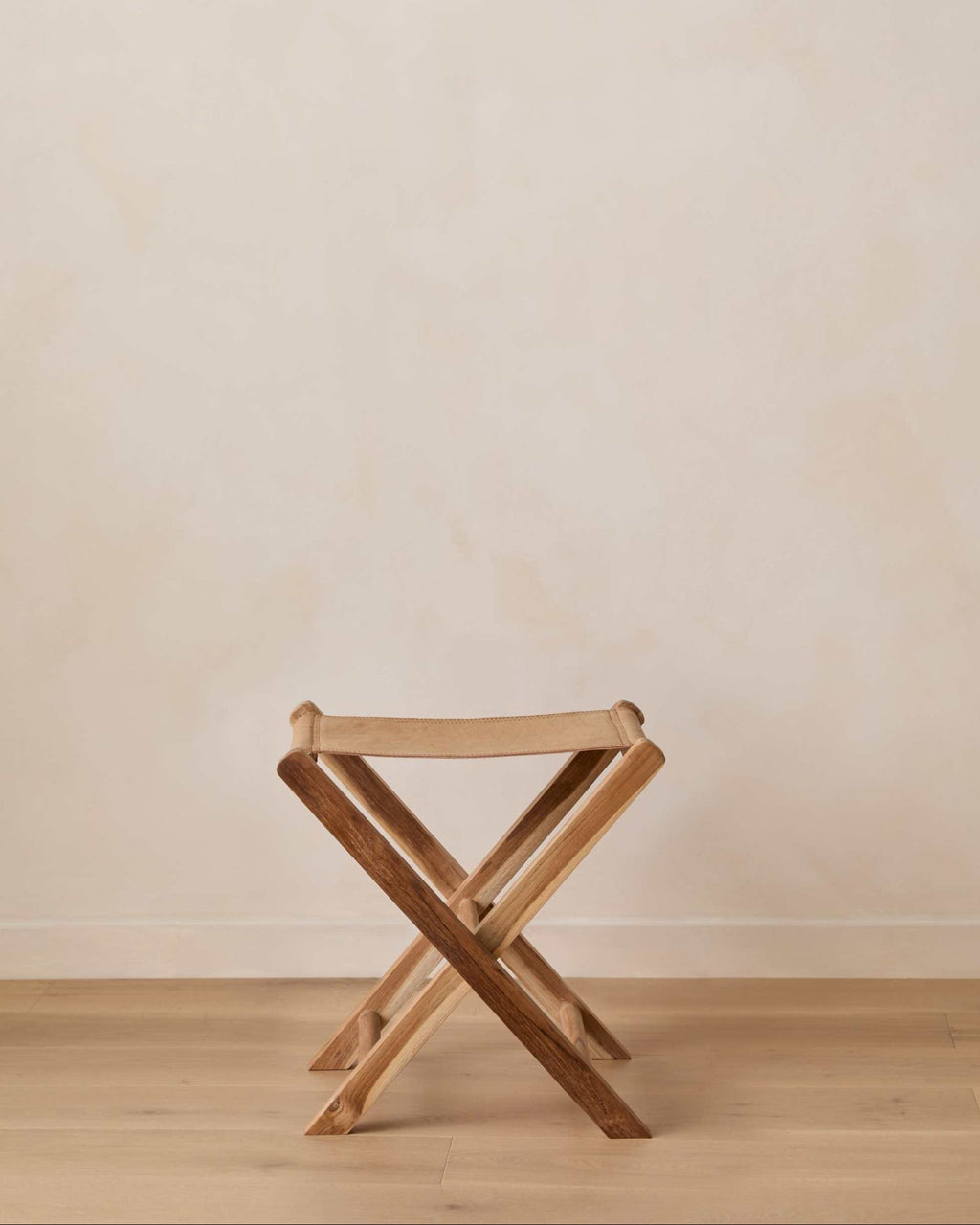 A small woven leather folding stool set against a warm beige wall and light wood floor.