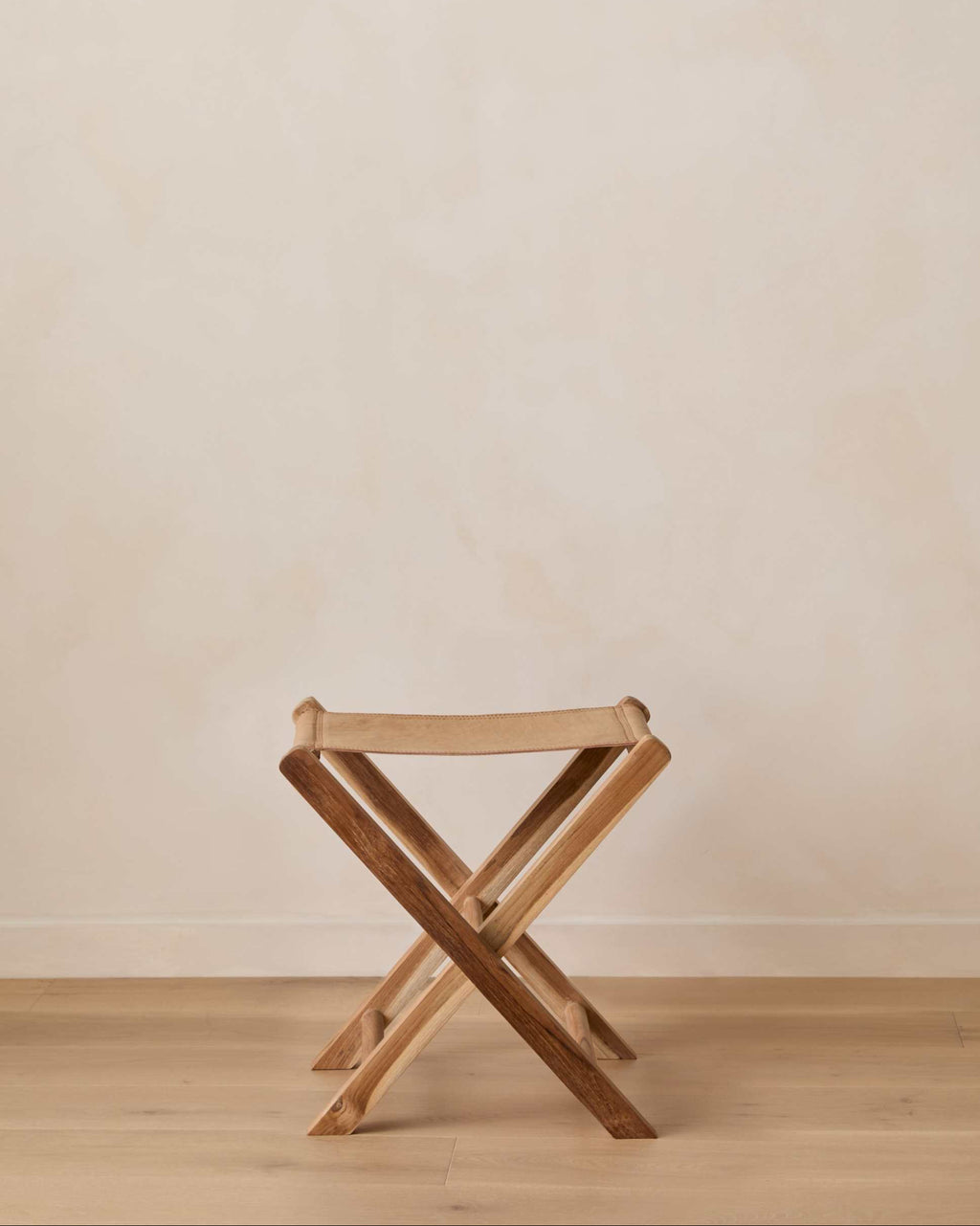 A small woven leather folding stool set against a warm beige wall and light wood floor.
