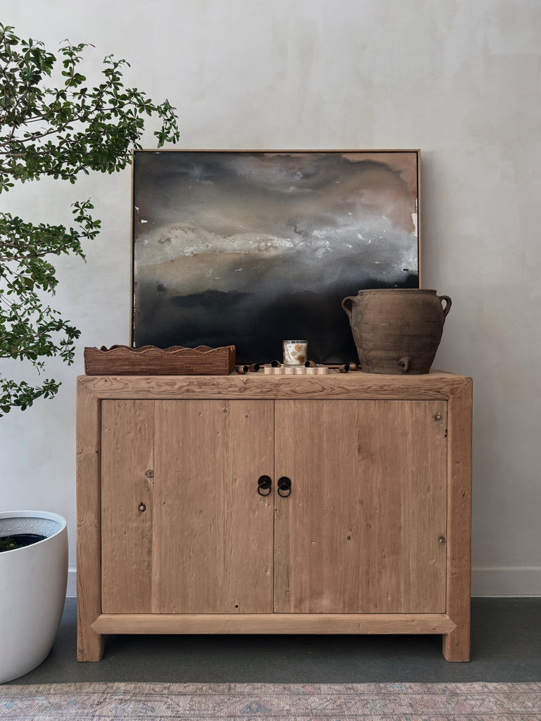 Wooden cabinet with decorative items and abstract painting against a limewashed wall