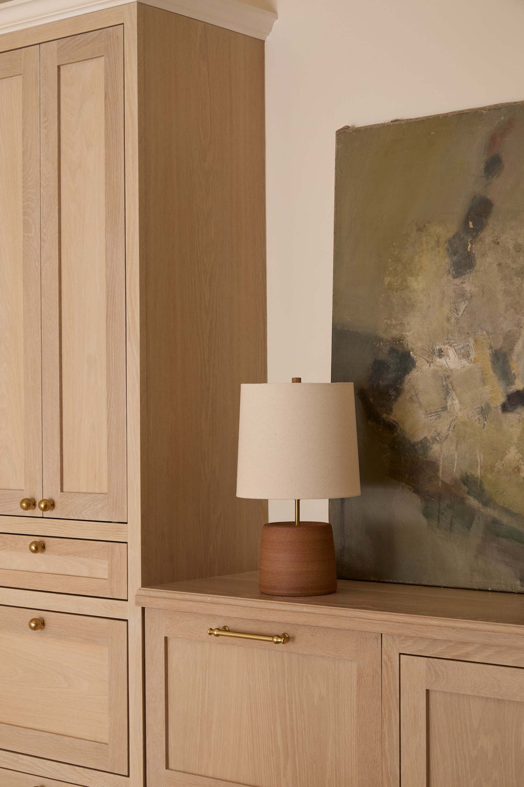 A light oak wood sideboard cabinet with brass handles and knobs, styled with a Ostuni terracotta table lamp with linen drum shade and a large abstract painting against a warm beige wall.
