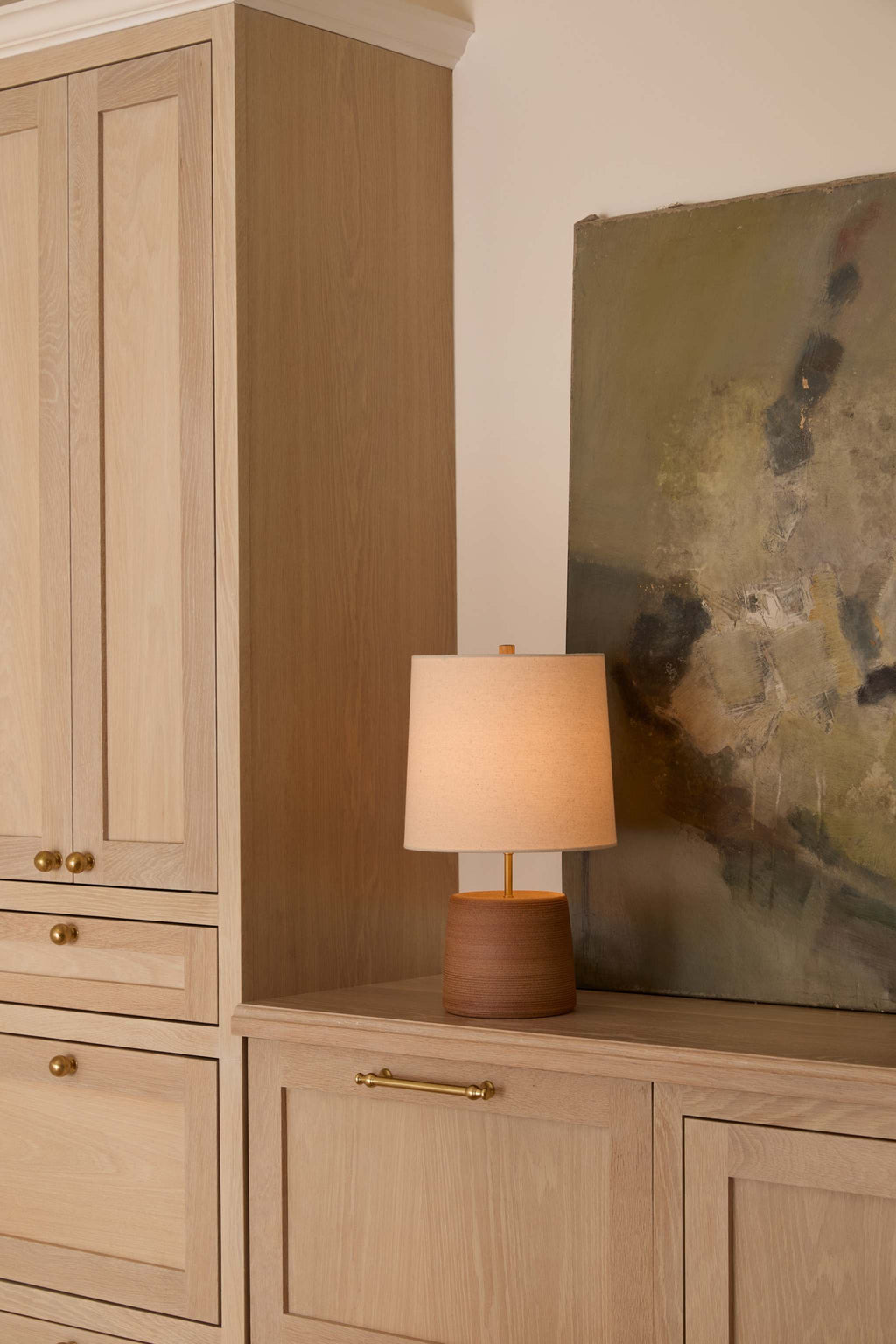 Ostuni table lamp with a warm beige linen fabric drum shade and a round textured terracotta base sits on a light oak credenza in a styled home interior next to a tall matching cabinet and an abstract painting.
