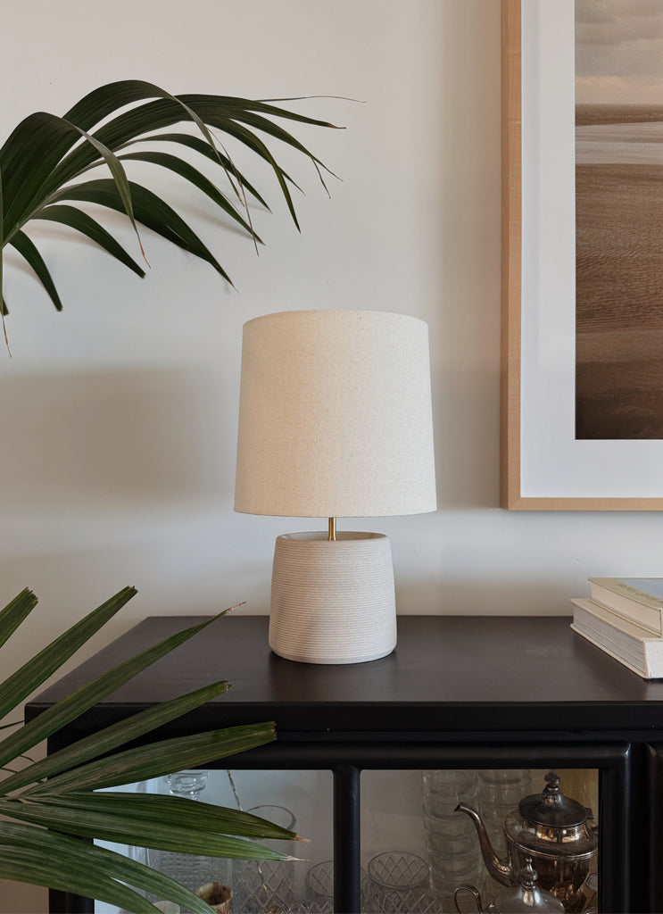 Ostuni Ceramic Table Lamp in Ivory with oatmeal linen shade on a black cabinet and plant. - Saffron + Poe