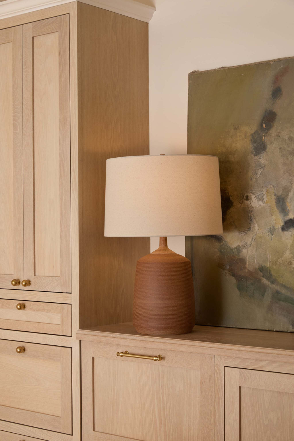 Pienza Ceramic table lamp with a textured terracotta base and a natural linen drum shade sits on a light wood cabinet in a styled interior setting.