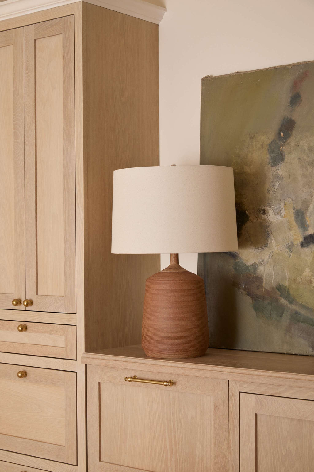 Pienza terracotta ceramic table lamp with a natural linen drum shade sits on a light wood credenza next to a large abstract painting.