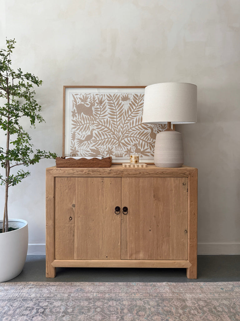 Pienza Ceramic Table Lamp in Ivory with oatmeal linen shade on a vintage wooden cabinet styled with decorative items. - Saffron + Poe