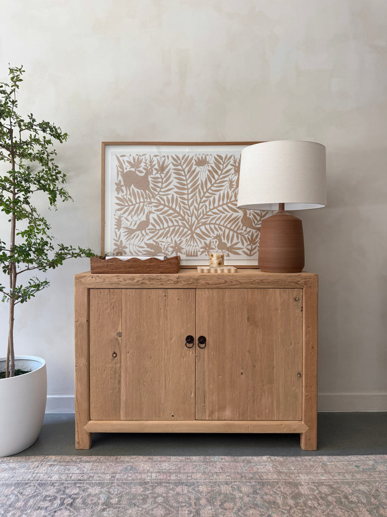 Pienza Ceramic Table Lamp in Terracotta with oatmeal linen shade on an antique cabinet styled with decorative items against a limewashed wall. - Saffron + Poe