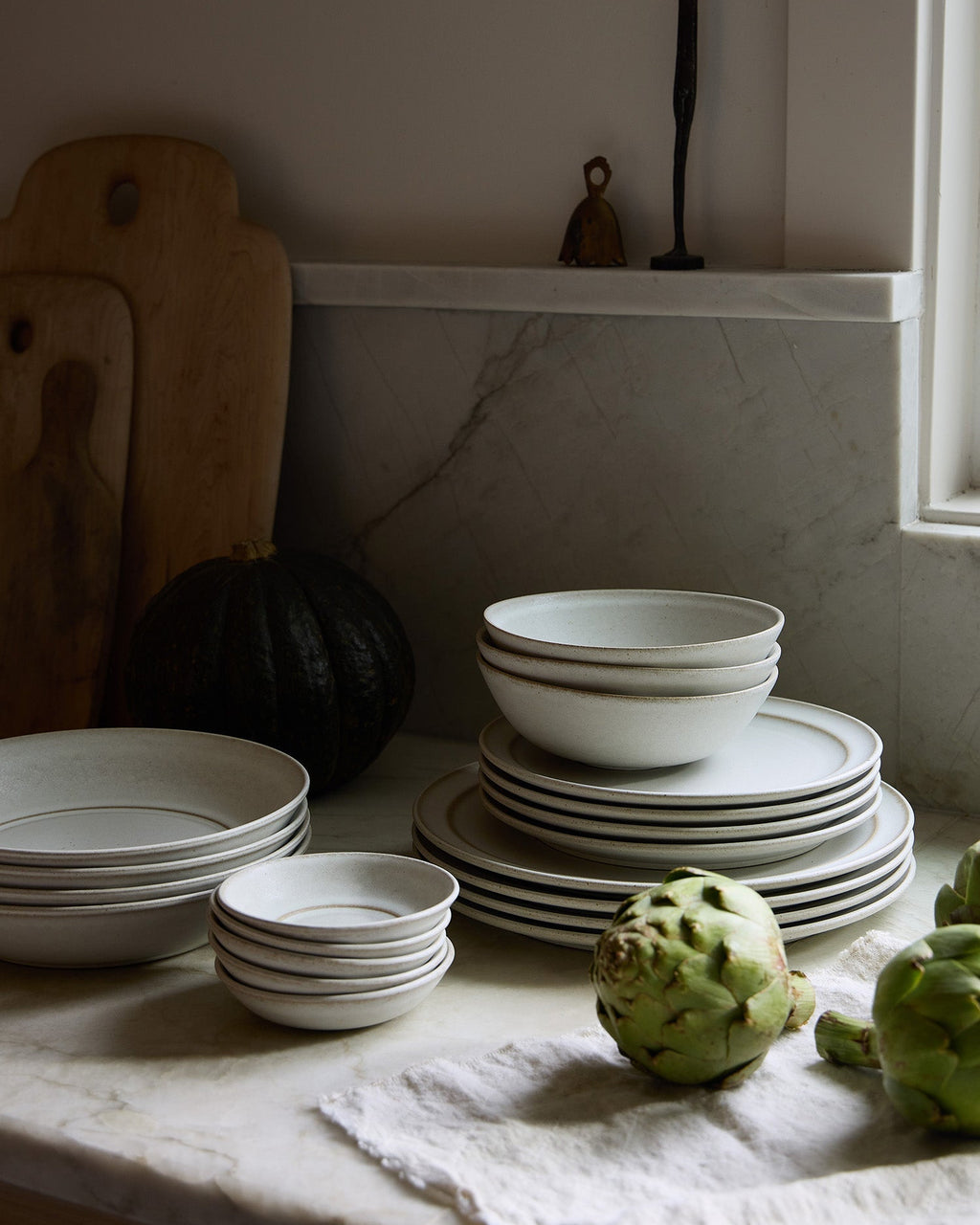 Stacks of white ceramic Pillar bowls and plates on a marble surface with a pumpkin and artichokes.