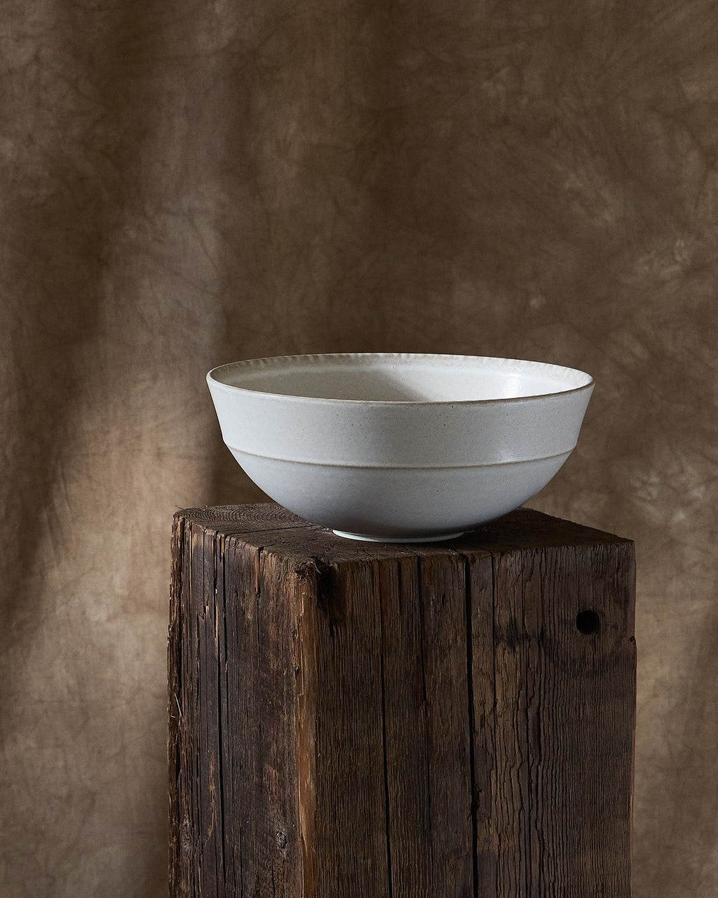 White ceramic Pillar Serving bowl on a wooden pedestal against a textured brown wall