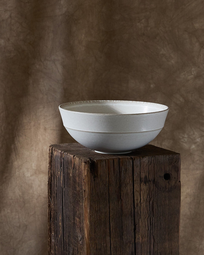 White ceramic Pillar Serving bowl on a wooden pedestal against a textured brown wall