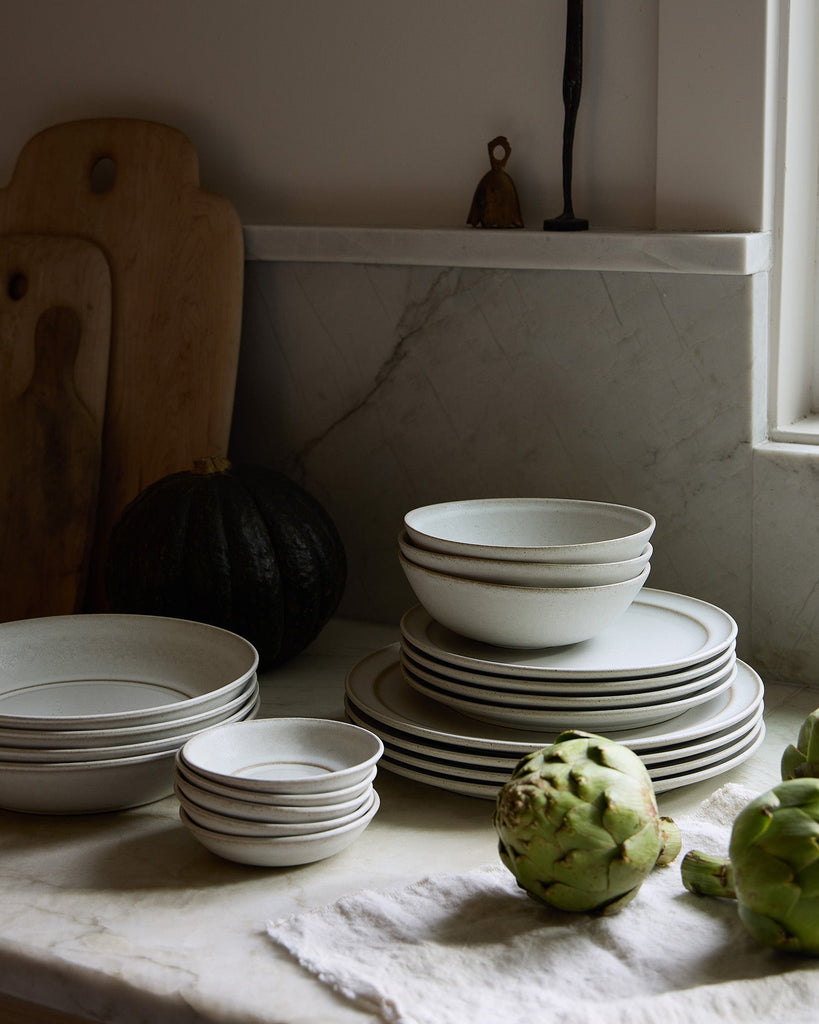 Stacks of white ceramic Pillar bowls and plates on a marble surface with a pumpkin and artichokes.