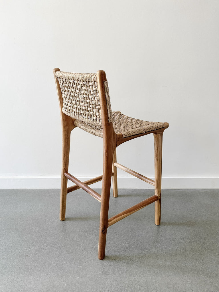 Back side product view of the Faux Rattan Outdoor Counter Stool on concrete flooring and a white wall backdrop. - Saffron and Poe