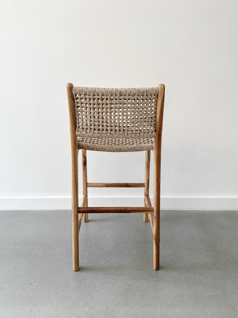 Back product view of the Faux Rattan Outdoor Counter Stool on concrete flooring and a white wall backdrop. - Saffron and Poe