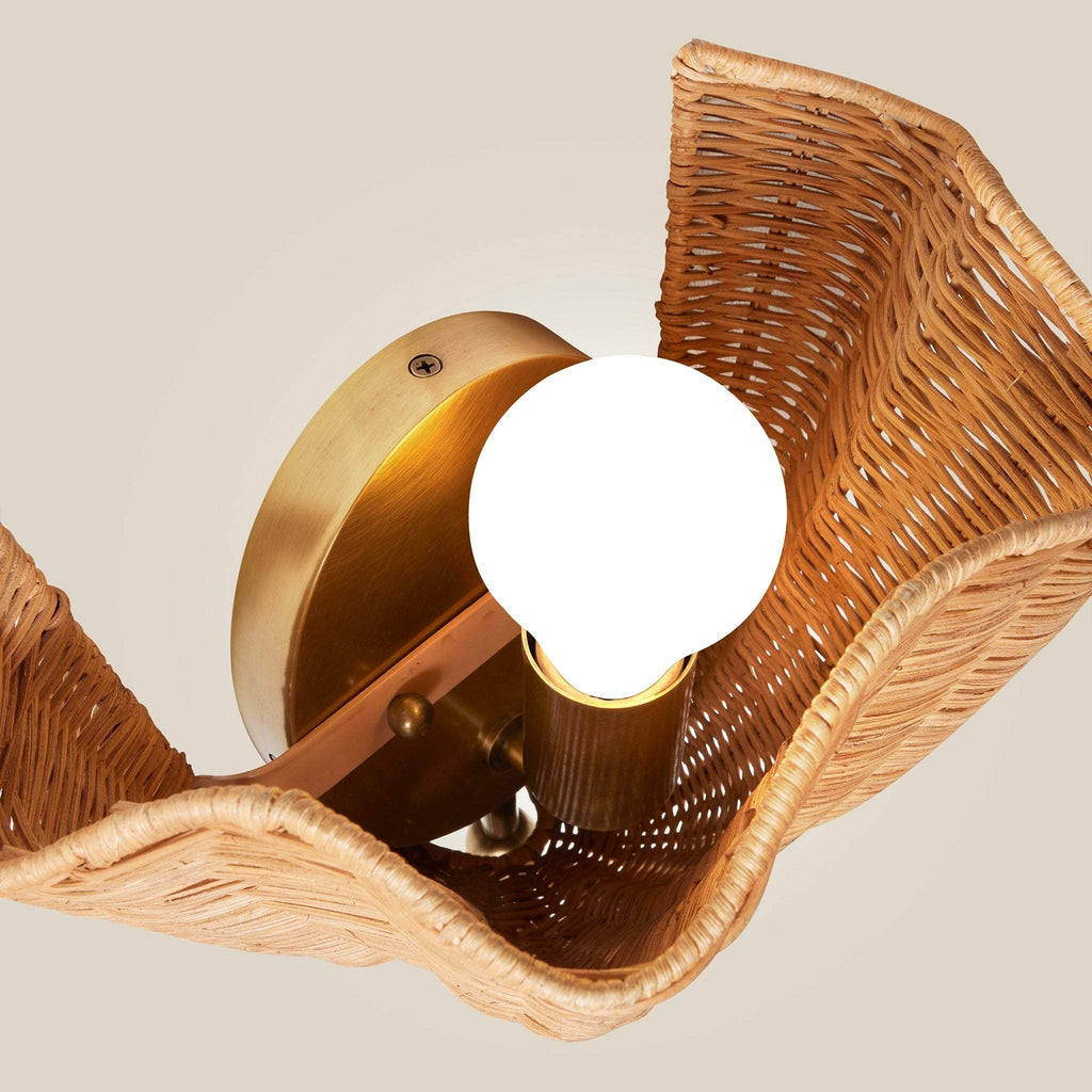 Close-up of a woven Ravello wall light fixture with a round white bulb and a warm brass metal base, shown against a plain light beige background.