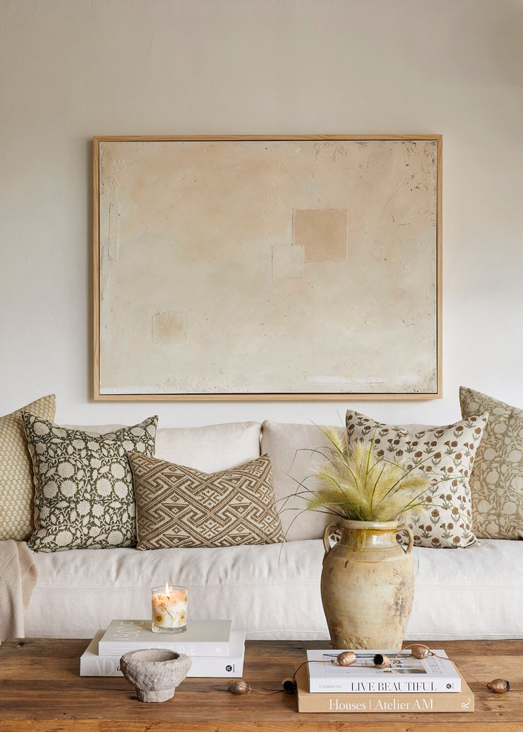 Living room with a white sofa, patterned throw pillows, a framed abstract painting, and decorative items on a wooden coffee table.