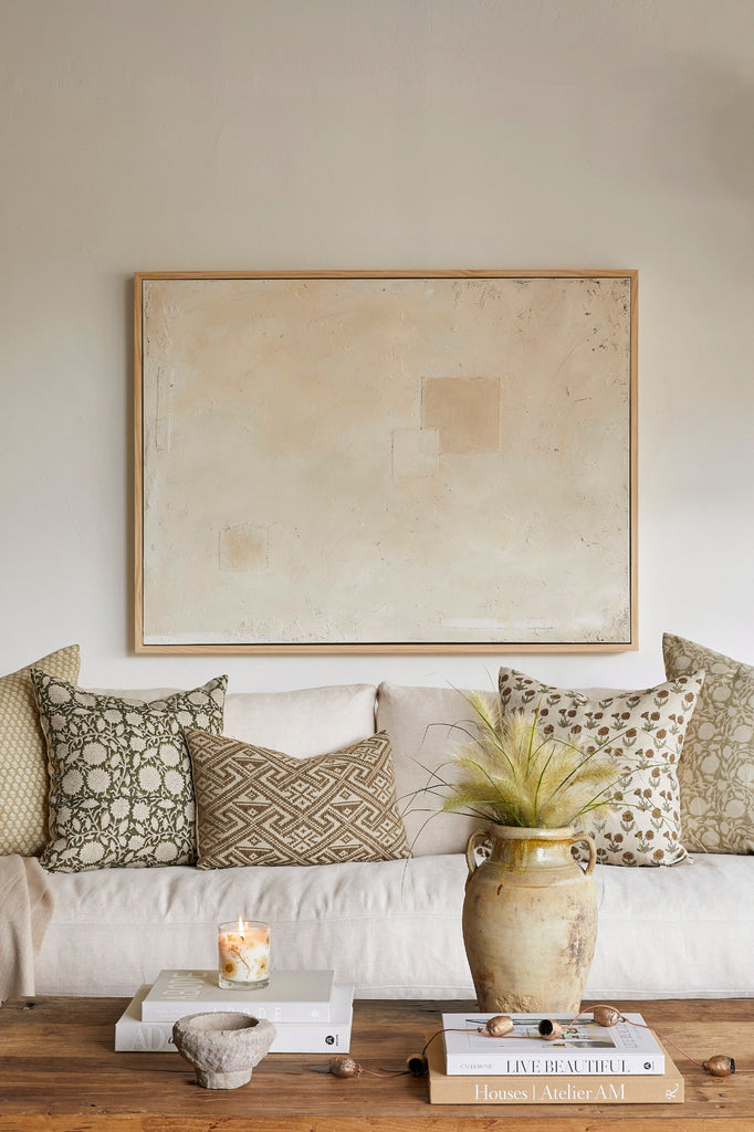 Living room with a white sofa, patterned cushions, a framed abstract painting, and decorative items on a wooden coffee table.