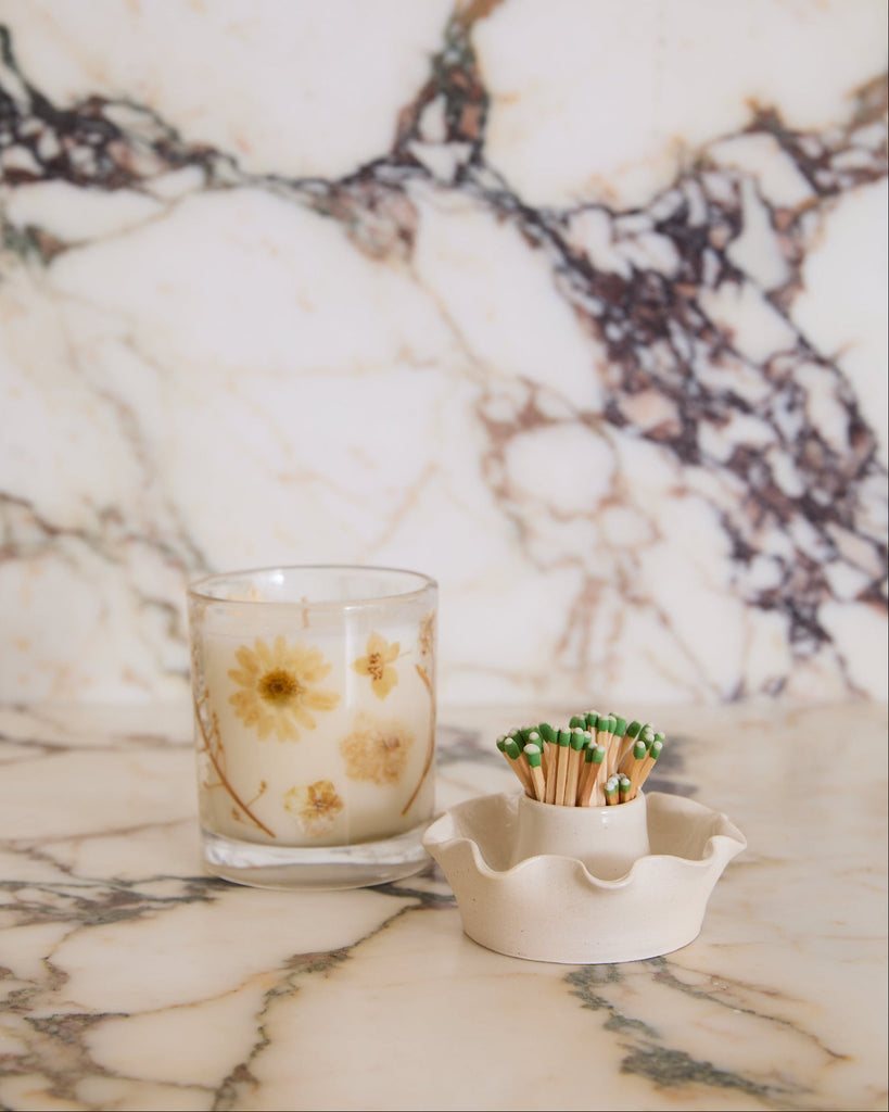 Scalloped ceramic match holder with candle with floral design on a marble surface.