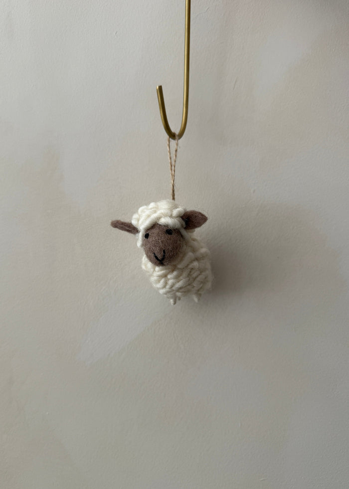 Knitted felt sheep ornament hanging on a hook against a lime washed wall. Saffron + Poe