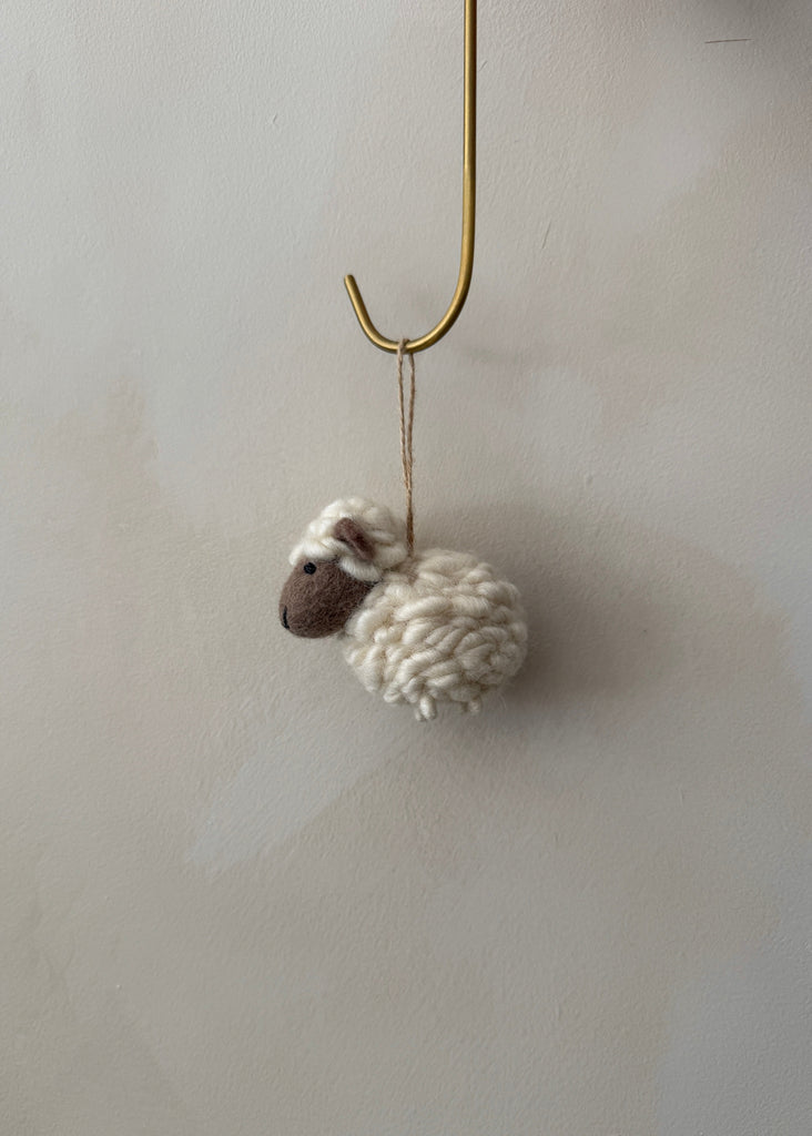 Knitted felt sheep ornament hanging on a hook against a lime washed wall. Saffron + Poe