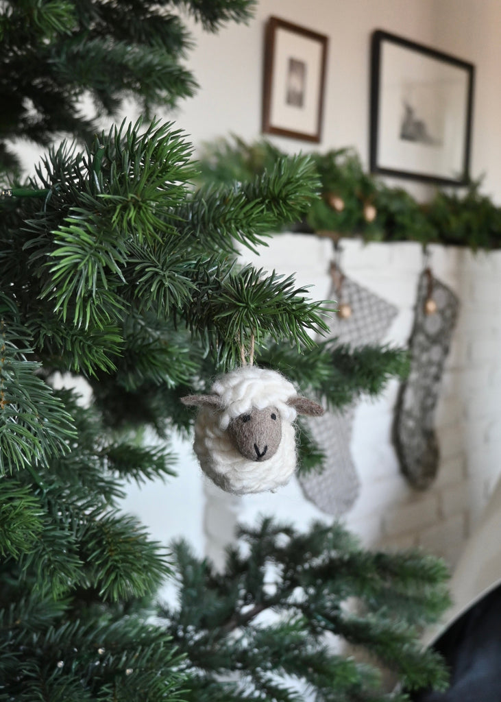 Knitted felt sheep ornament hanging on a Christmas tree with stockings in the background. Saffron + Poe