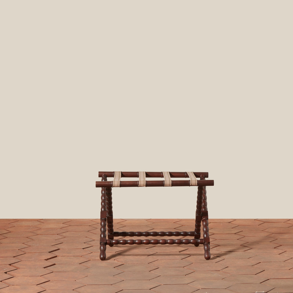 Dark brown mahogany wooden folding luggage rack with bobbin legs and a woven beige and brown cotton straps set on a terracotta tiled floor against a solid beige wall.