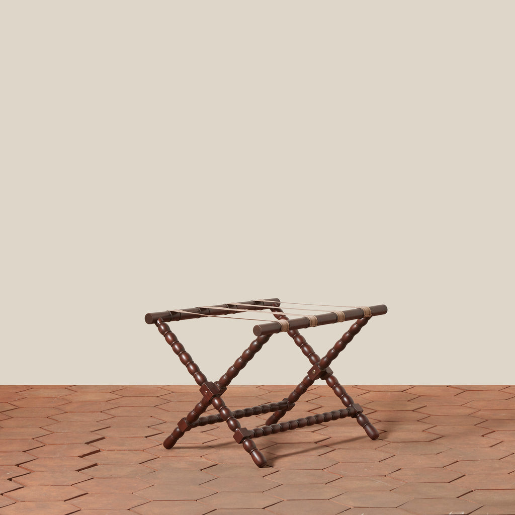Foldable dark brown mahogany wood Somerset Luggage Rack with a spoked leg design and cotton straps, positioned on a terracotta tiled floor against a solid beige wall.