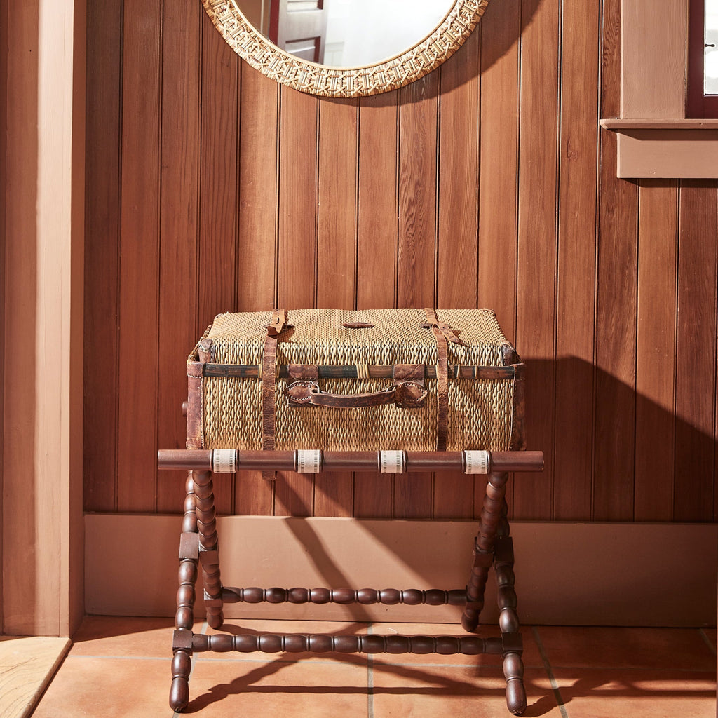 Mahogany Somerset Luggage Rack with bobbin legs and beige cotton straps styled with a vintage-style woven suitcase with leather straps against a warm wood-paneled wall.