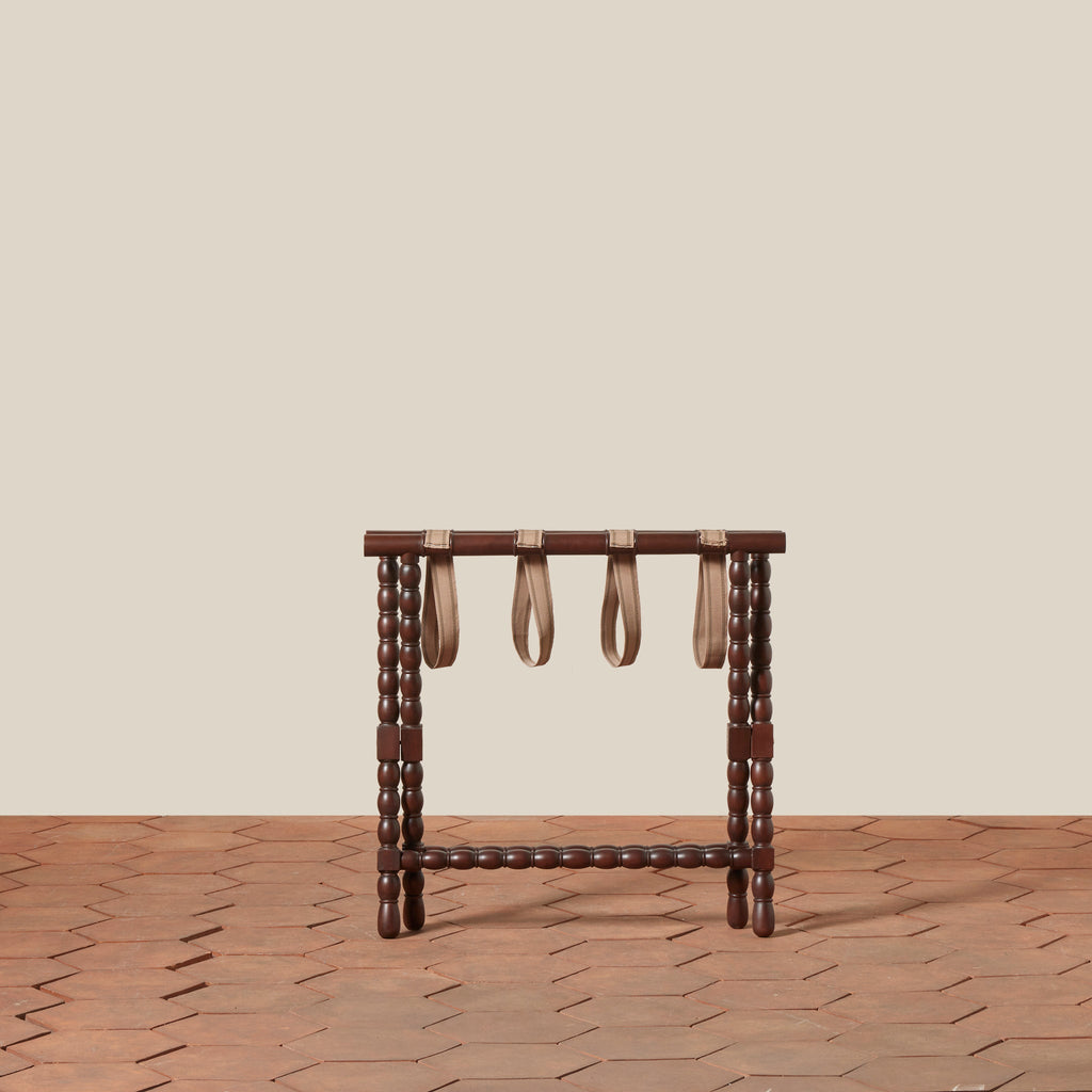 Somerset Luggage Rack with mahogany bobbin legs and four beige cotton straps, set against a minimalist beige wall and a terracotta hexagonal tile floor.