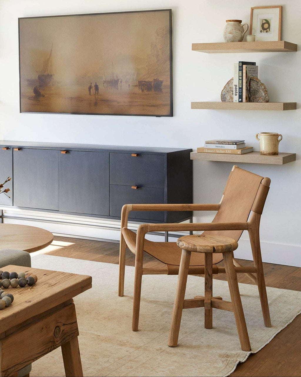 A modern living room scene featuring a mid-century wooden armchair with a woven seat, a vintage elm wood stool, a dark gray sideboard, and a large abstract landscape painting on the wall.