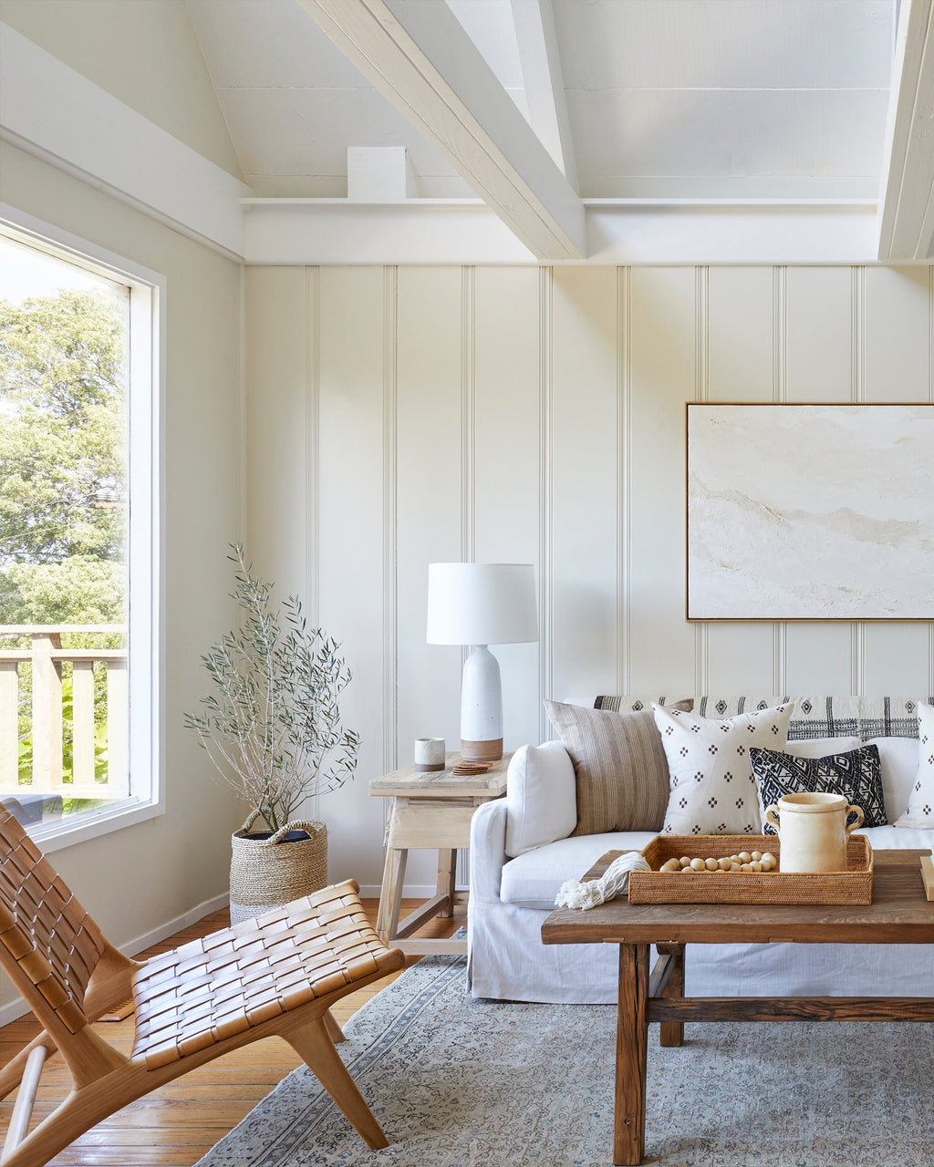 A bright, modern living room featuring a white sofa with neutral and patterned pillows, a wooden coffee table, a woven leather lounge chair, and a large framed abstract print on a cream paneled wall.