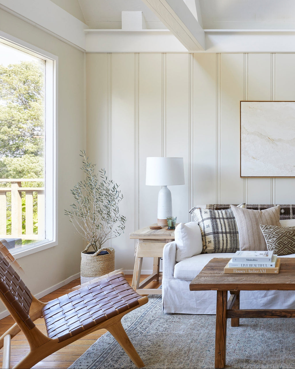 A bright, modern living room with white paneled walls and ceiling beams, featuring a woven leather lounge chair, white sofa with patterned pillows, a wooden coffee table, and a large framed abstract print on the wall.