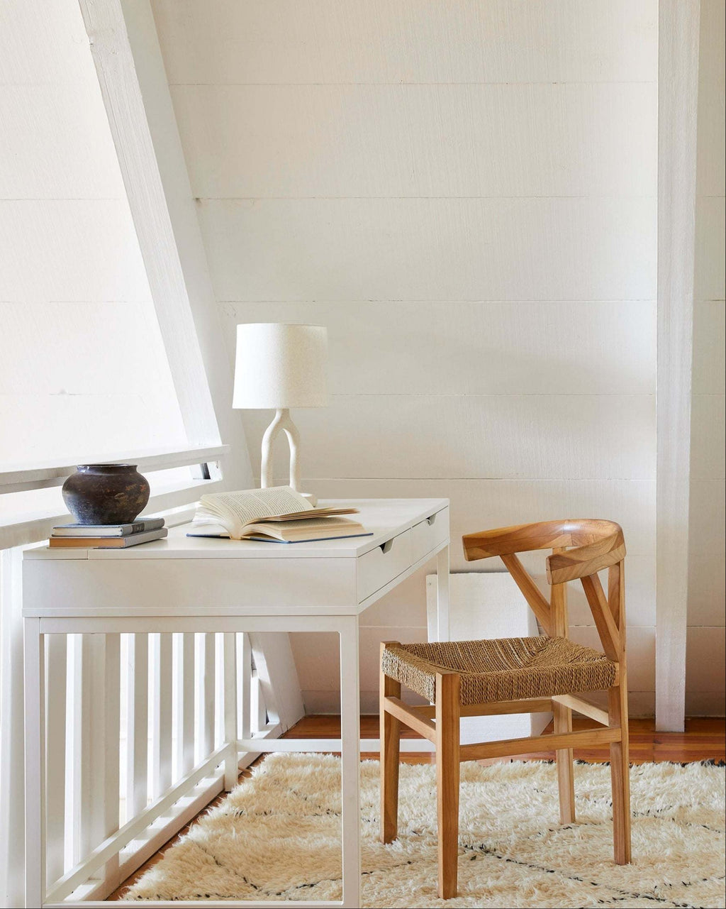 A white writing desk with a matching white lamp and an open book, styled with a woven wishbone dining chair with a woven seat on a cream shag rug in a bright room with white paneled walls.