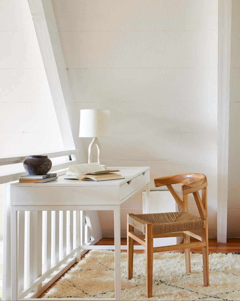 A white writing desk with a matching white lamp and an open book, styled next to a woven wishbone dining chair with a woven seat on a cream shag rug in a bright room with white paneled walls.