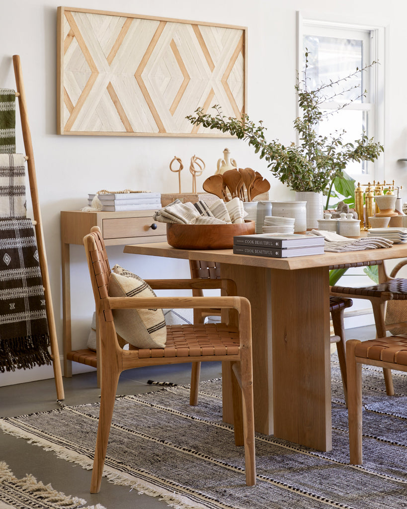 Styled side view of the Woven Leather Strap Dining Arm Chair in Beige in a dining room setting with Aleksandra Zee art work, Oak Dining Table, and Moroccan Rug.