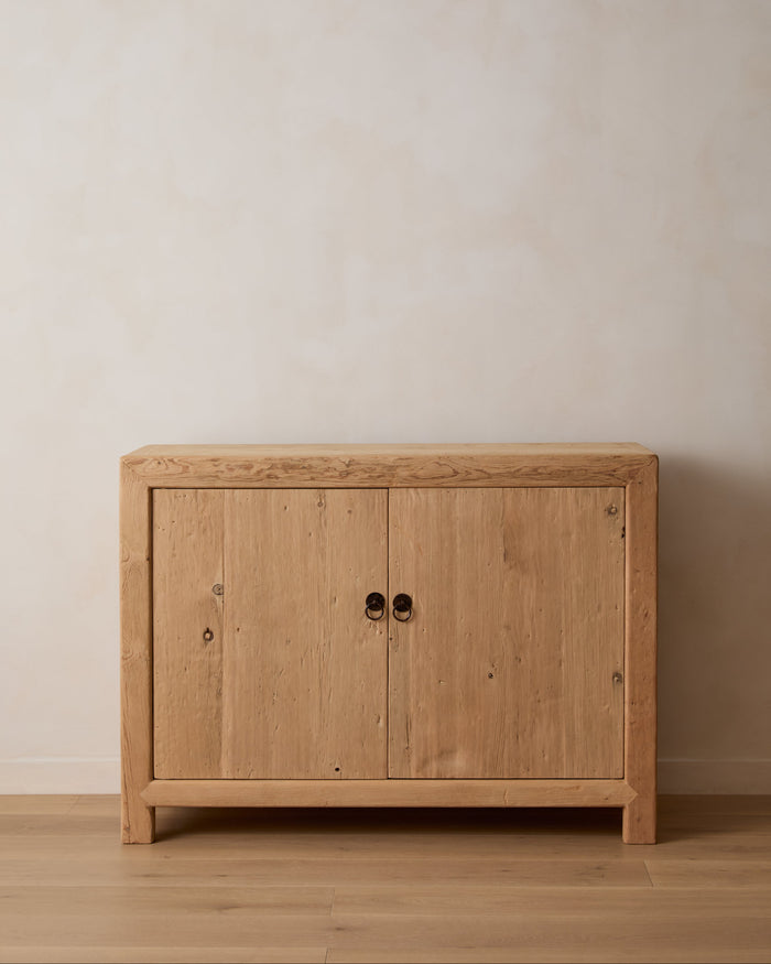 A Vintage light natural Elm wood sideboard cabinet with two doors and round black metal handles, set against a neutral beige wall and light wood floor.
