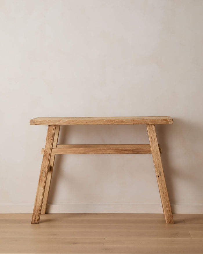 A light natural vintage elmwood console with two angled legs connected by a lower bar, set against a plain beige wall and light wood floor.