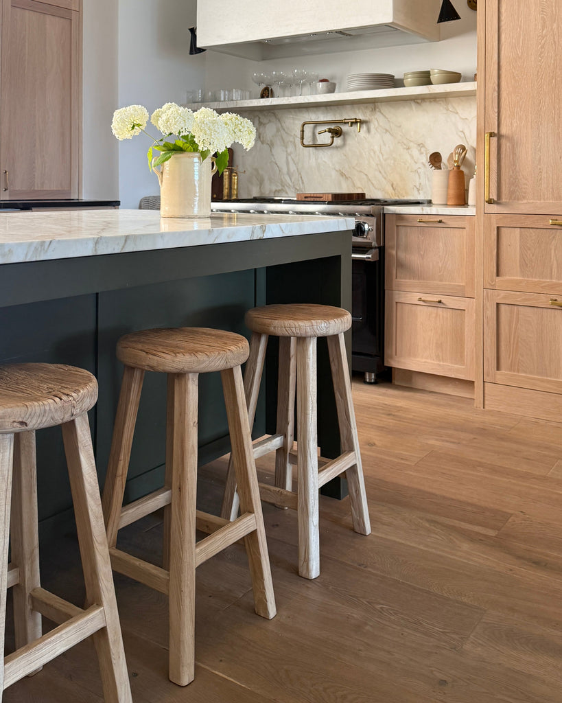 Modern kitchen with vintage elmwood counter stools at an island, light wood cabinets, and marble countertop. 