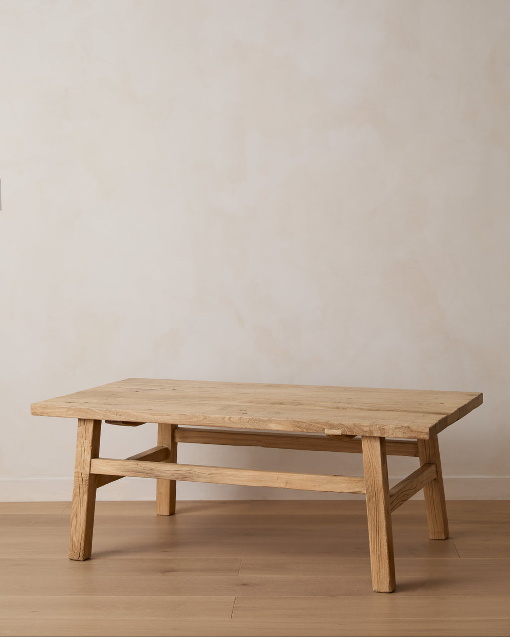 Vintage wood rectangular coffee table in light elm with a thick top and four legs, shown in a minimalist room setting with a textured beige wall and light wood floor.