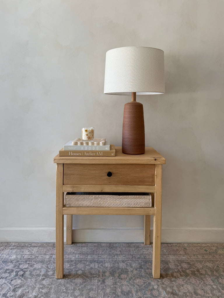 Vintage Reclaimed Elmwood Bedside Table with hammered drawer knob styled with ceramic lamp in front of beige limewashed wall. Saffron + Poe