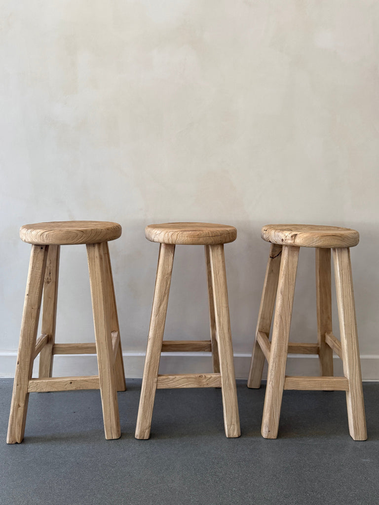 Three Vintage Reclaimed Elmwood Counter Stool in a row against beige limewashed wall. Saffron + Poe