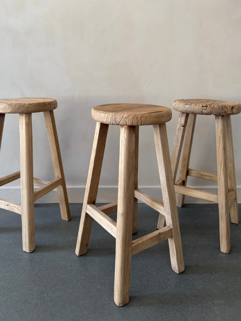 Three Vintage Reclaimed Elmwood Counter Stool grouped against beige limewashed wall. Saffron + Poe