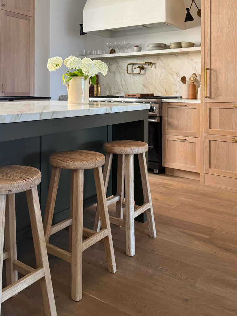 Modern kitchen with vintage elmwood counter stools at an island, light wood cabinets, and marble countertop. Saffron + Poe