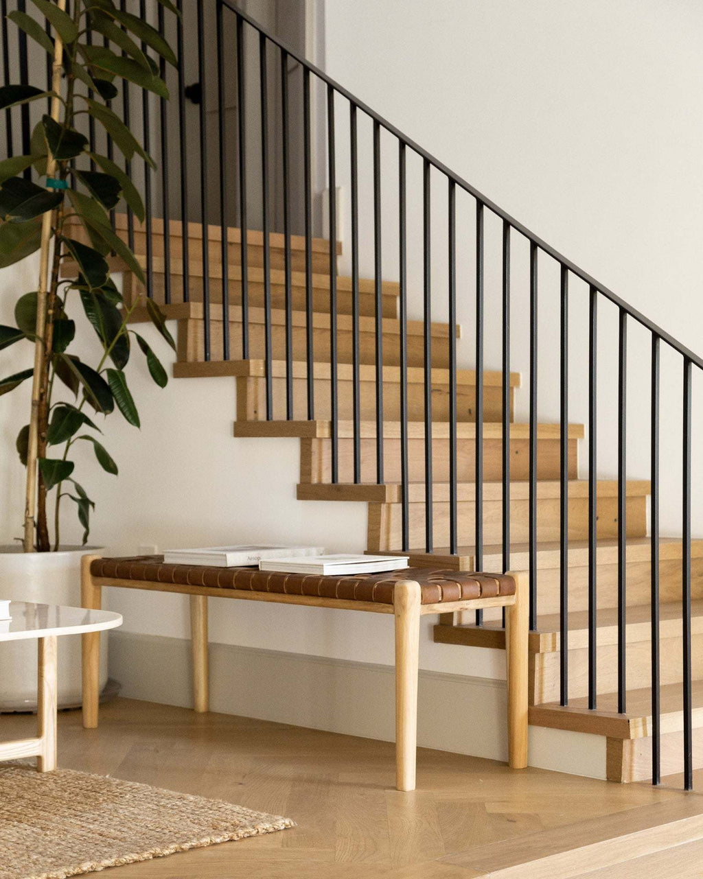A modern wooden staircase with black metal balusters, featuring a saddle woven leather bench in the landing.