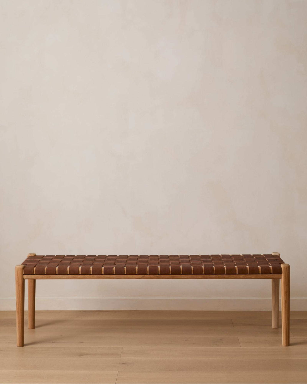 A modern wooden bench with saddle leather woven seat, set against a minimalist beige wall and light wood floor.