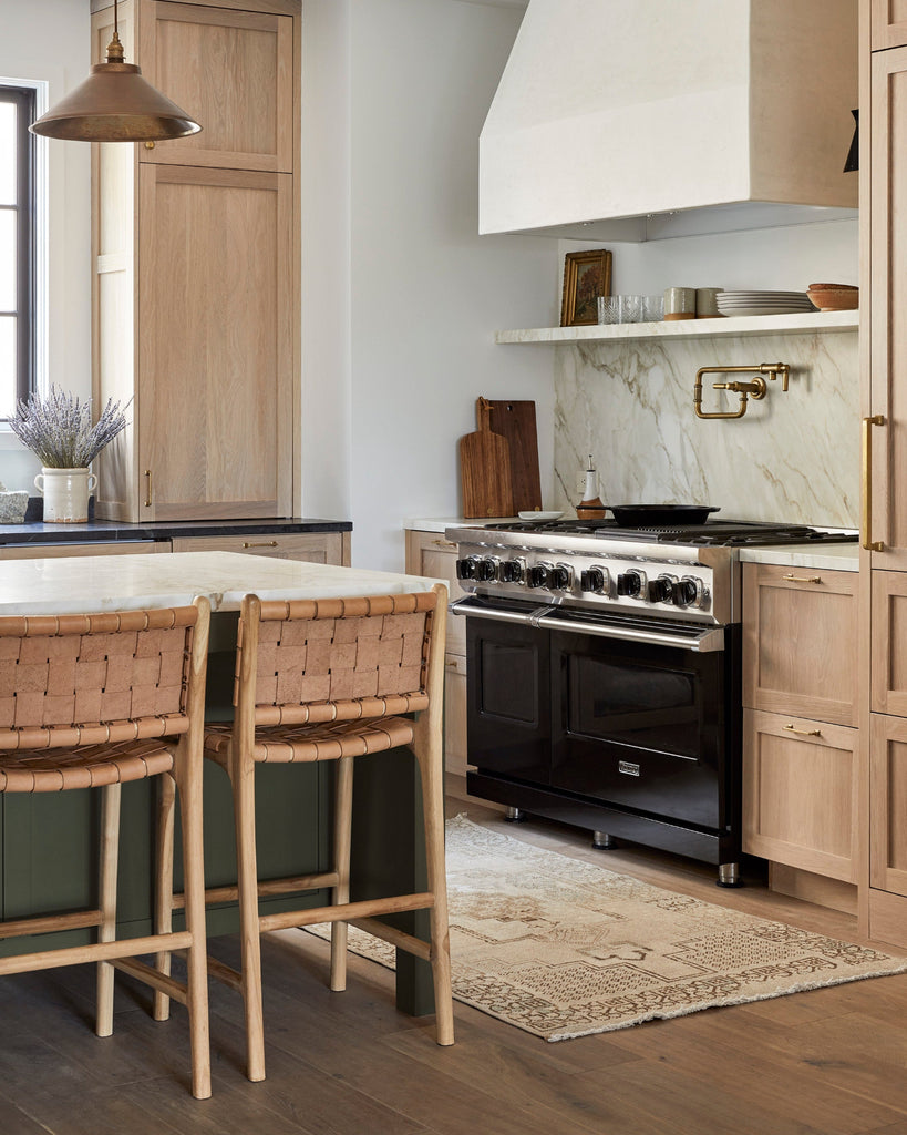 Modern kitchen with black stove, beige woven leather counter stools, wooden cabinets, and marble backsplash.